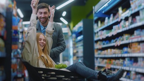 Happy young family couple carries a girl in a grocery basket shopping in supermarket. Smiling lovely husband and wife buyers customer grocery store concept. Slow motion