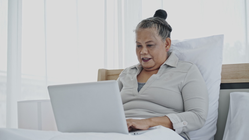 Asian senior woman sitting on a laptop Or happy notebook On white bed in the house or hotel. Finding Healthcare Information, Shopping Online And video conference. Tele consultation with a doctor | Shutterstock HD Video #1063181185