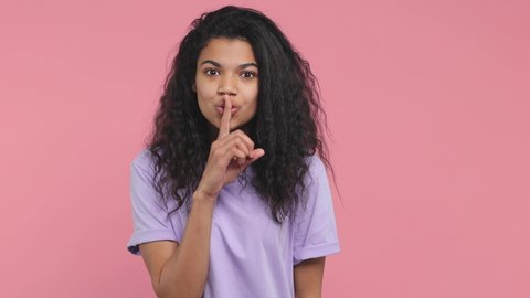 Secret young african american woman in casual violet t-shirt isolated on pink background in studio. People lifestyle concept. Looking aside say hush be quiet with finger on lips shhh gesture blinking