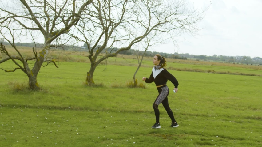 Strong young woman runs up hill in a field by trees