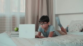 Home Isolation, Online Learning, Remote Work, New Education. Boy doing homework using online technologies while lying on the bed at home