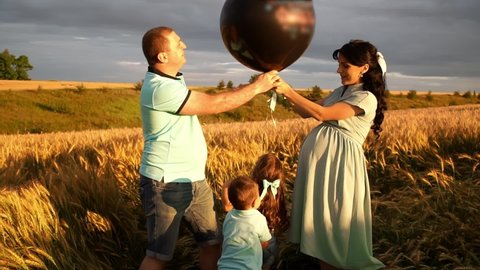 Family with two children standing in field pops air balloon to know gender of future baby in slowmo. All members are happy that there will be a girl. They are smiling and hugging. Reveal party theme.
