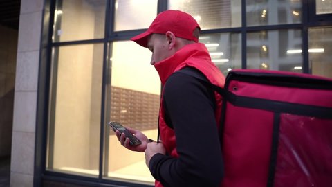 Delivery guy wearing red uniform while walking along modern buildings down city street with thermal bag. Close up follow Shot