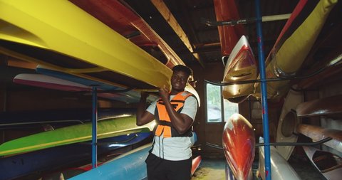 Happy young African American male kayaker holding canoe while standing in warehouse with many colorful kayaks. Handsome teen guy in life vest indoors smiling in good mood. Summer sport concept Arkivvideo