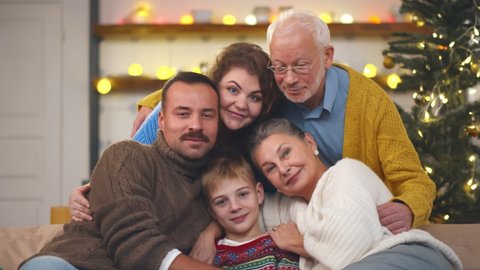 Portrait of large family embracing and smiling at camera with christmas tree on background. Happy grandparents, parents and cute boy hugging and looking at camera in decorated apartment