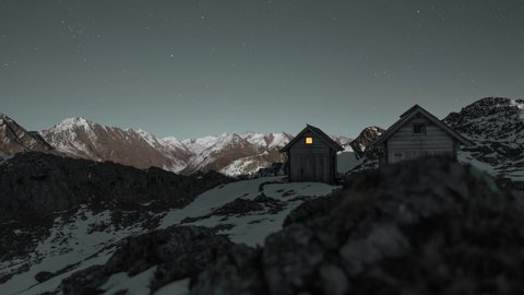 Two mountain huts in the Alps. In one of the huts there are people staying overnight and the window is illuminated. Timelapse withing moonlight and stars.