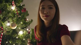 Young Asia female drinking beer having fun happy night party video call talk with couple, Christmas tree decorated with ornaments in living room at home. Christmas night and New Year holiday festival.