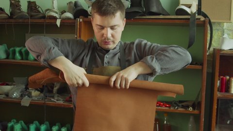 A shoemaker's apprentice rolls a piece of genuine leather into a roll. In the background, racks with blanks for shoes.