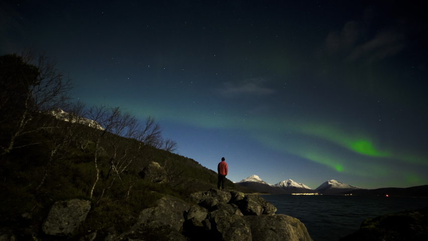 Man standing by the rocks facing the lake as he enjoys the beautiful scenery and skies with Northern Lights