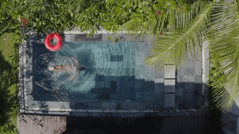 SLOW MOTION: Young woman diving inside private swimming pool. Top down aerial view young female dives in slow motion into blue clear water 