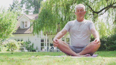 Senior man at home in garden wearing fitness clothing sitting cross legged on yoga mat and meditating - shot in slow motion