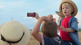 smiling man holds smartphone in his hand and takes selfie on camera in phone, with young woman and boys on background of haystacks while walking in field with bales of hay outsid