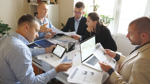 Business people discussing income charts and graphs during team meeting in modern office. Young colleagues sitting at the table and analyzing financial reports. Coworkers examining documents at desk