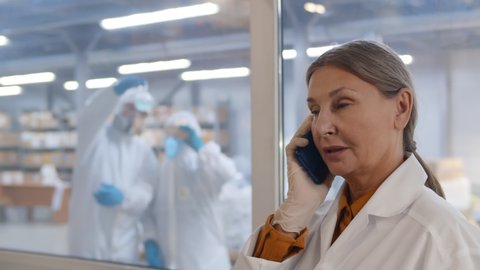 Mature woman scientist in white coat and gloves talking on phone at modern chemical factory. Female laboratory worker having phone call with colleagues in overalls carrying experiment behind window
