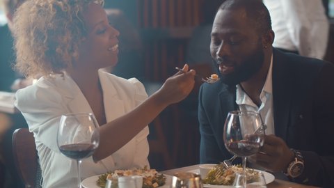 Loving black couple having dinner date feeding each other sitting in restaurant. Portrait of happy elegant african man and woman drinking wine and dining in modern restaurant