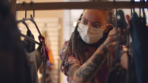 Young woman wearing safety mask choosing clothes in rack in shop. Hipster female customer in protective face mask shopping clothes in retail shopping store.