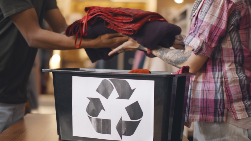 Close up of volunteers packing used clothes in container with recycling sign. Man donating old clothing in charity shop for recycling and reuse. Royalty-Free Stock Footage #1063233166