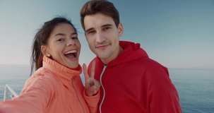 Happy young couple in tracksuits standing on background of sea shooting video on smartphone. Beautiful girl kisses guy on cheek.
