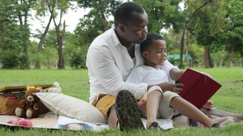 Low angle shot of a happy African family outing at park concept. Outdoors, medium shot of dark skin, kind father and a son picnic at the park, sitting on the mat, and reading a book together.
