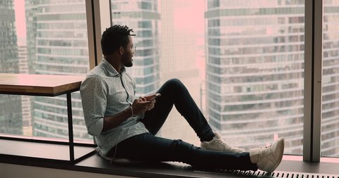Full length relaxed african guy, communicate remotely using smartphone admires city view, sit on windowsill in modern apartment or office room near panoramic window with skyscrapers cityscape scene