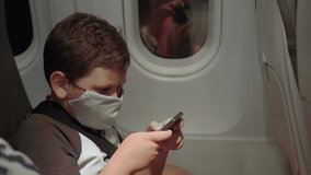 Boy in protective face mask to protect against the coronavirus pandemic plays video games on the phone on an airplane while flying at night
