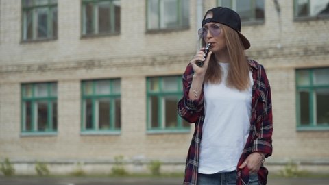 Pretty young hipster woman in black cap vape ecig. Girl vape popular ecig gadget, vaping device.Happy female vaper with e-cig. Portrait of smoker model with electronic cigarette vaporizer. Slow motion