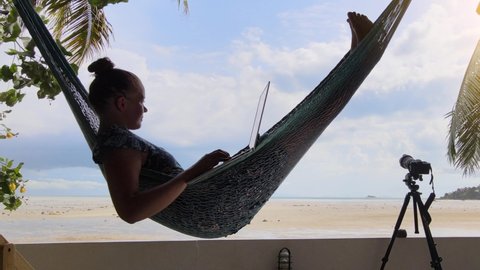 Digital nomad girl remote working on a laptop in the hammock on the tropical island beach house. Camera on tripod. Palm trees and a sandy beach in the background. Content creator or influencer job