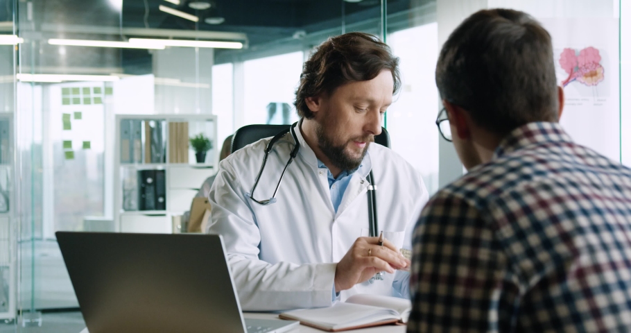 Back view of male patient sitting at clinic in cabinet on consultation with doctor. Close up portrait of handsome man physician at work speaking with patient and showing medicine. Healthcare concept | Shutterstock HD Video #1063251928