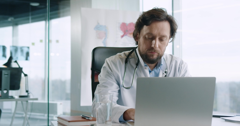 Portrait of handsome adult busy Caucasian male doctor sitting at desk in hospital office working and typing on laptop at workplace. Healthcare worker tapping on computer. Job concept | Shutterstock HD Video #1063251958