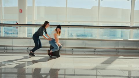 Two friends fool around, run and make jokes in airport. Friend push woman on top of suitcase, joking around in empty airport terminal, wait for delayed or cancelled flight. Teenagers travelling