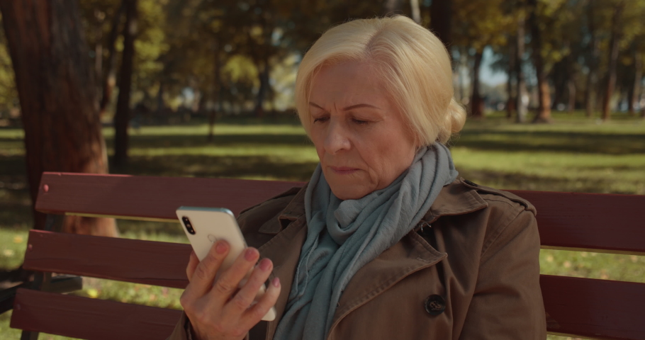 Worried senior female reading shocking news on smartphone, sitting in park