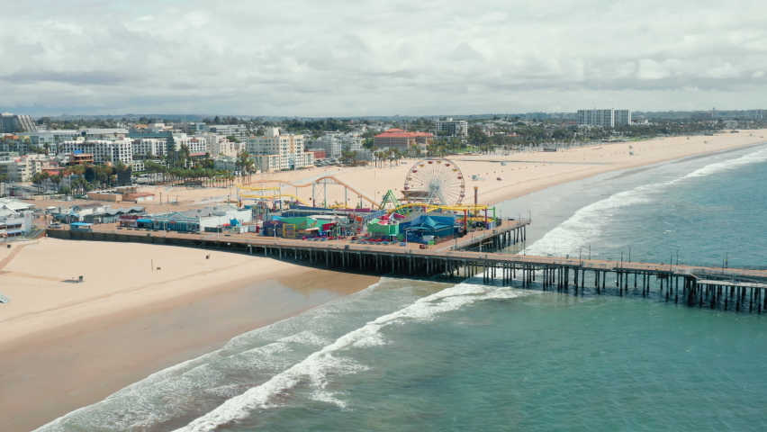 Santa Monica Pier on cloudy day, Los Angeles. The footage of the Santa Monica pier and the amusement park from the side of the ocean. Beautiful panorama of the city skyline is seen behind sandy beach Royalty-Free Stock Footage #1063262560