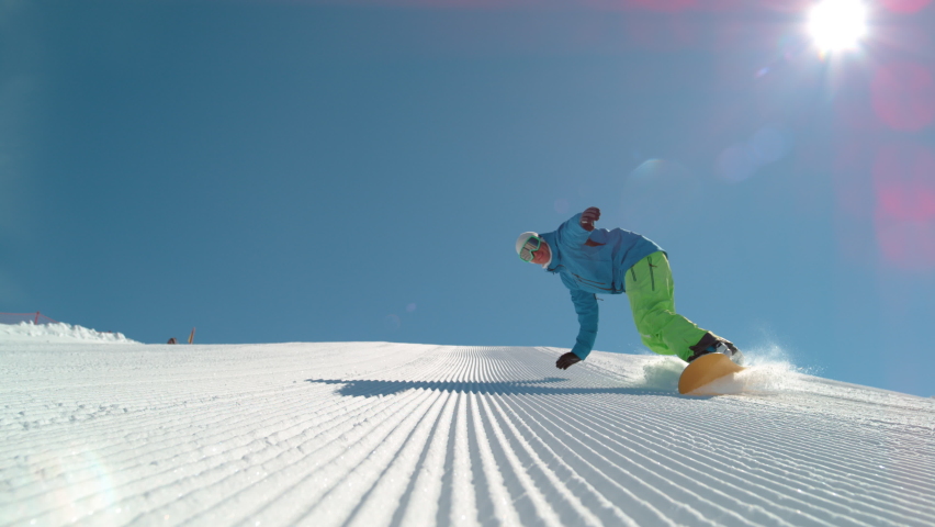 SLOW MOTION TIME WARP, LOW ANGLE, CLOSE UP, DOF: Athletic male tourist shredding the slopes sprays snow at the camera. Snowboarder carves along a groomed ski slope of a ski resort in the sunny Alps. Royalty-Free Stock Footage #1063265098