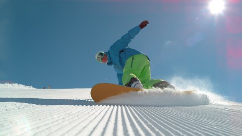 SLOW MOTION TIME WARP, LOW ANGLE, CLOSE UP, DOF: Athletic male tourist shredding the slopes sprays snow at the camera. Snowboarder carves along a groomed ski slope of a ski resort in the sunny Alps.