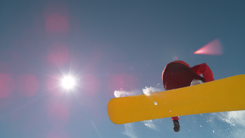 SLOW MOTION TIME WARP, LENS FLARE, BOTTOM UP: Fearless snowboarder does a spin trick while riding off a big air kicker. Snowflakes sparkle in the sunshine as male tourist does snowboarding tricks. Royalty-Free Stock Footage #1063265116