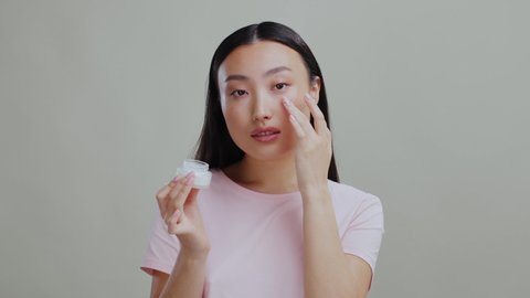 Healthy looking korean woman applies moisturizing day cream holding ba jar posing isolated on grey studio background. Skincare routine.