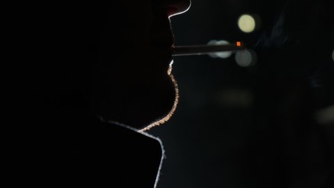 Close up view of smoking man rack focus at night in park with lights in the background. man with bristles smokes cigarette and blows smoke out of his mouth.