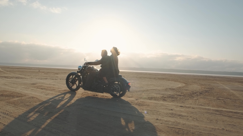 Guy with a girl on a motorcycle in the desert. Couple riding on vintage motorcycle and having a good time at sunset on a dry salt lake. Slow motion shot Royalty-Free Stock Footage #1063268956