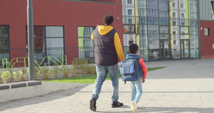 Rear of happy African American male parent walking on street while bringing little cute son to school. Loving father with junior shool student with backpack walk to school outdoor. Knowledge concept Royalty-Free Stock Footage #1063269205