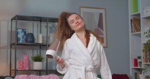 Close up of young blonde woman with long hair wearing white bathrobe drying hair with hair dryer after shower singing and dancing in living room. Concept of lifestyle, self-care and wellness.