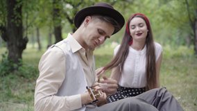 Smiling elegant young man playing ukulele with blurred beautiful woman listening to serenade at the background. Side view portrait of loving Caucasian boyfriend dating with girlfriend on picnic. - Powered by Shutterstock - Get 15% off with code: PIKWIZARD15