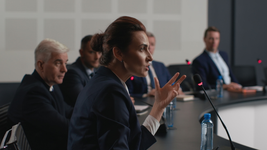 Political speaker talks at meeting room of modern business forum. Adult woman in suit discusses partnership at conference in convention hall. Expert group of colleagues works at official event closeup Royalty-Free Stock Footage #1063272478