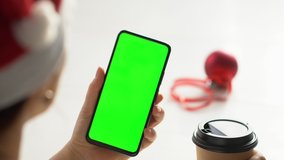 Close-up shot of green screen template smartphone in female hands at home office. The template can be used to advertise purchases on Christmas Eve. Modern technology and information concept. POV
