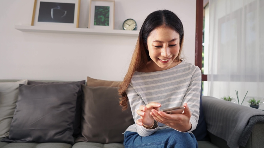 30s young adult Asian woman playing a mobile phone sits on sofa in the living room at home. Happy female internet and technology user in casual clothes on couch in cozy room - 4K Slow Motion Footage
