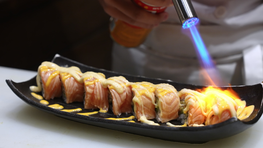 Close up of Professional chef hand burning salmon sushi roll with shrimp tempura and mayonnaise on serving plate. Chef preparing healthy sushi menu on open kitchen counter bar in japanese restaurant. Royalty-Free Stock Footage #1063283518