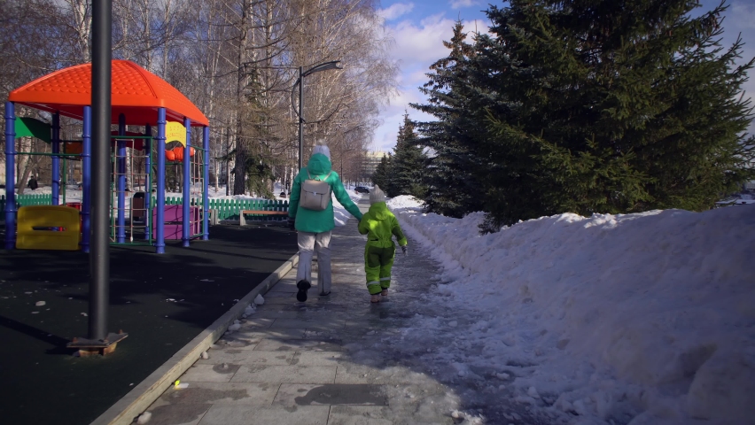 A small child and a mother together holding hands walk on a wet sidewalk next to a playground and snowdrifts, a view from the back.