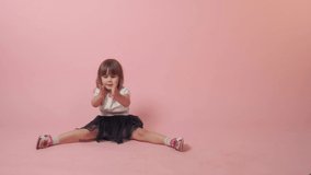 A cute little girl sits on the floor and shows her ears with her hands, laughs and indulges. Beautiful bright festive outfit. Pink background.