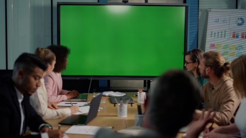 Multi ethnic business team looking at presentation on big tv with green screen in office. Business people having online video conference during quarantine. Coworkers working in meeting room together