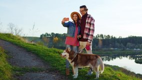 Portrait attractive young couple use phone and take selfie have video call stand with dog in the forest near lake. Nature mobile relationship. Outdoors. Romantic. Close up. Slow motion