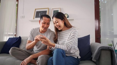 30s young adult Asian man and woman playing a mobile phone together sits on sofa in cozy living room at home. Happy couple internet user in casual clothes on couch. Technology usage in Asia concept